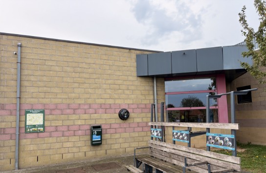Bushalte met houten bankje en informatieborden naast een gebouw met gele en roze bakstenen. (vergroot foto)