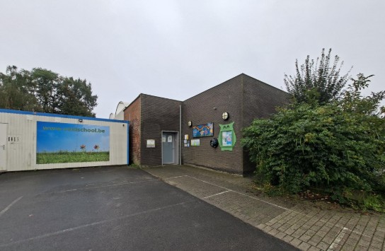Ingang van een schoolgebouw met een parkeerplaats en groene struiken, onder een bewolkte lucht. (vergroot foto)
