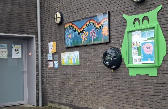 Muur met kleurrijke borden, een groene uil-vormige prikbordkast en een AED kast bij een gebouwingang. (vergroot foto)