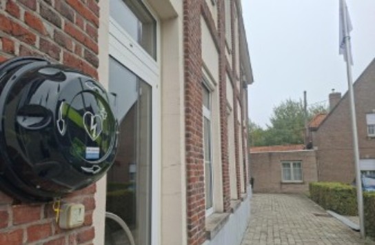 Elektrische laadpaal aan gevel van bakstenen gebouw met deur en straatje ernaast op bewolkte dag. (vergroot foto)