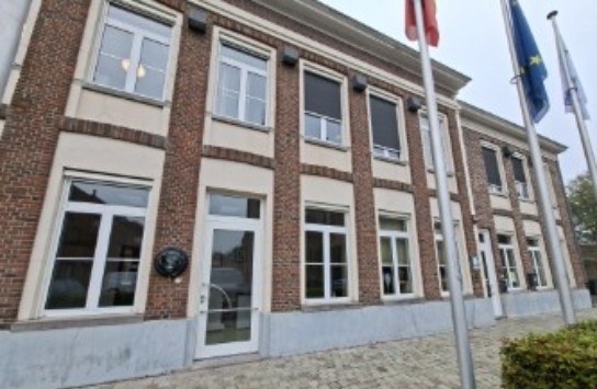 Vooraanzicht van een klassiek gebouw met bakstenen gevel en meerdere vlaggenmasten ervoor. (vergroot foto)