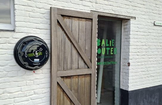 Houten deur half open naast een witte bakstenen muur met een AED kast en groene tekst op glas. (vergroot foto)