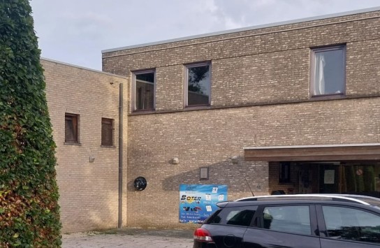 Parkeerplaats met zwarte auto voor een modern gebouw van beige baksteen en een groene heg links. (vergroot foto)