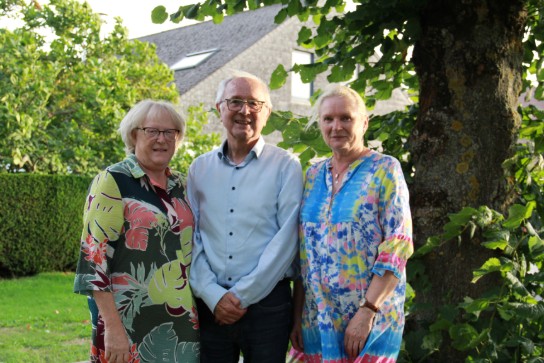 Drie volwassenen staan buiten in een tuin, omringd door groen en een boom, bij een huis met een rieten dak. (vergroot foto)
