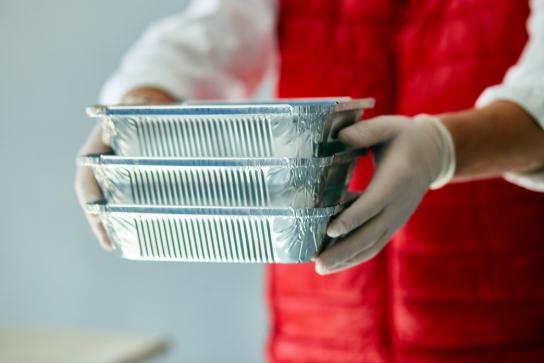 Persoon met witte handschoenen en rood vest houdt drie gestapelde aluminium voedselbakjes vast. (vergroot foto)